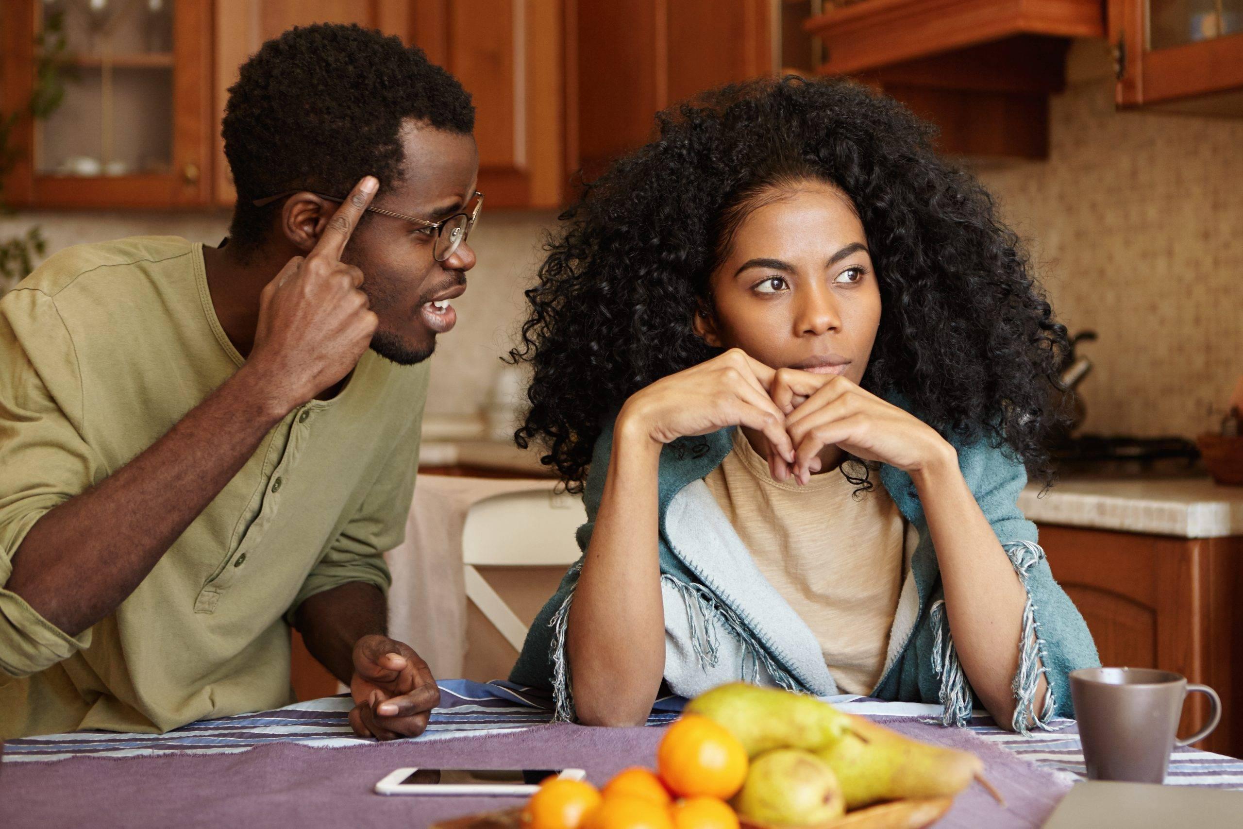 Couple having dispute. Annoyed beautiful dark-skinned female sitting at kitchen table, ignoring screams and insults from her mad furious husband who is shouting at her, holding finger at his temple
